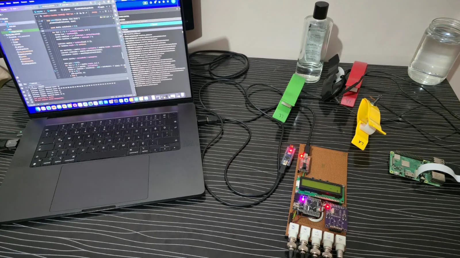 The live system during a development session: application source on the laptop, the board powered with its display active, limb clips and the alcohol and saline prep staged